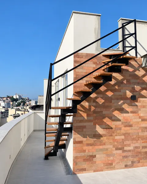 Escada externa de viga central preta com degraus de madeira junto à parede de tijolo aparente, sob céu azul